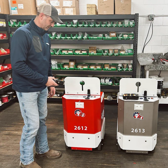A person stands in an industrial setting beside two small robotic machines labeled "2612" in red and "2613" in gray. The person is holding a device, possibly controlling the robots. Shelving with various parts and boxes is visible in the background.