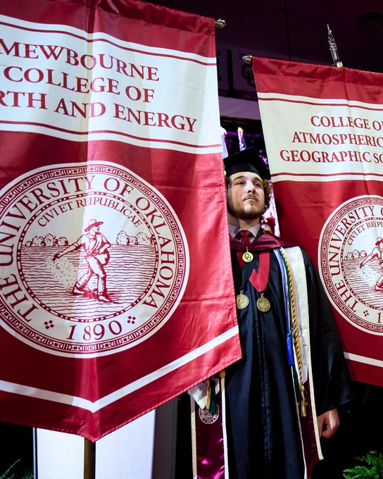 A person in academic regalia stands between two University of Oklahoma banners. The banners read "Mewbourne College of Earth and Energy" and "College of Atmospheric & Geographic Sciences," each featuring the university's seal.