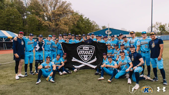 Kent State baseball team photo taken on the field at their home ballpark on an overcast day. The full roster poses together in their light blue "Flashes" alternate jerseys with navy and gold trim, holding up a large black pirate-style flag featuring the MAC (Mid-American Conference) logo with "1946" above it and crossed baseball bats beneath. Players are arranged in two rows — standing in the back and kneeling in the front — all smiling and celebrating. The outfield fence, a blue team facility building, trees, and a "320" distance marker are visible in the background. The bottom right corner features the Kent State Ohio logo, the Golden Flashes "K" emblem, and the Under Armour logo.
