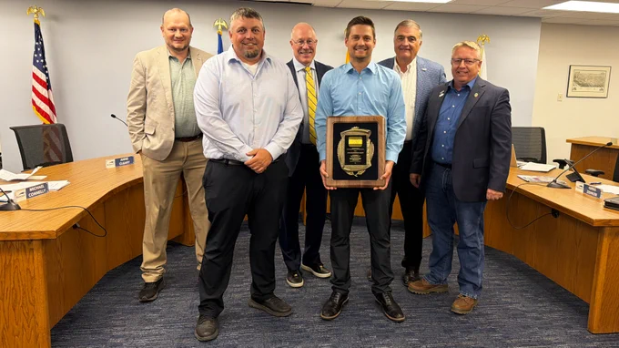 A group of individuals standing together in a formal meeting room, with one person holding a large award plaque.