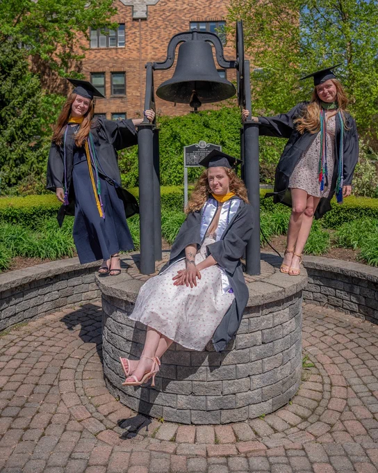 Three students posed by the bell in Trinity Green, dressed in their caps and gowns. 