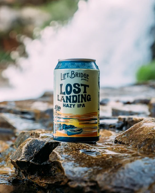 A can of Lift Bridge Brewing Co.'s Lost Landing Hazy IPA is placed on wet stones in front of a blurred waterfall. The setting evokes a natural and refreshing atmosphere.