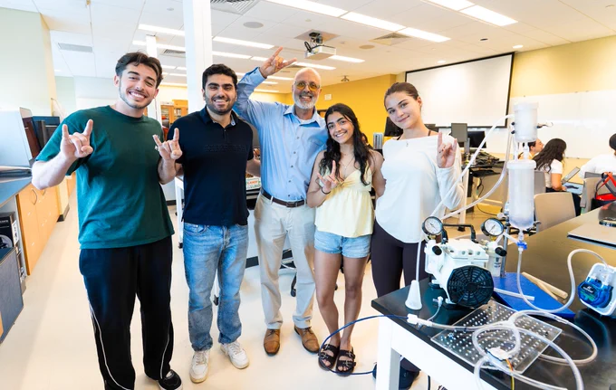 A group of five people stands smiling in a bright laboratory, all making the "Horns Up" hand sign. There is scientific equipment on a table nearby, including a device with dials and tubes. The room has a projector screen and desks in the background.