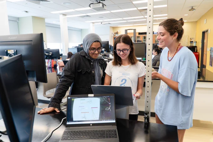 A group of three individuals in a lab setting are gathered around a laptop on a desk. There are computers and technical equipment visible in the background. The room is bright and orderly, with large windows allowing natural light.