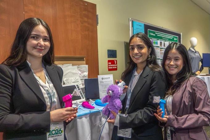 Three people stand smiling in front of a display booth. They are dressed in business attire and wear badges. The background features informational posters and a mannequin.