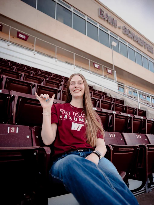 A person is seated in a sports stadium, wearing a maroon "West Texas A&M Alumni" shirt. They're making a hand gesture. The seating area features several numbered maroon seats, and the background shows a building with "Bain-Schaeffer" on it.