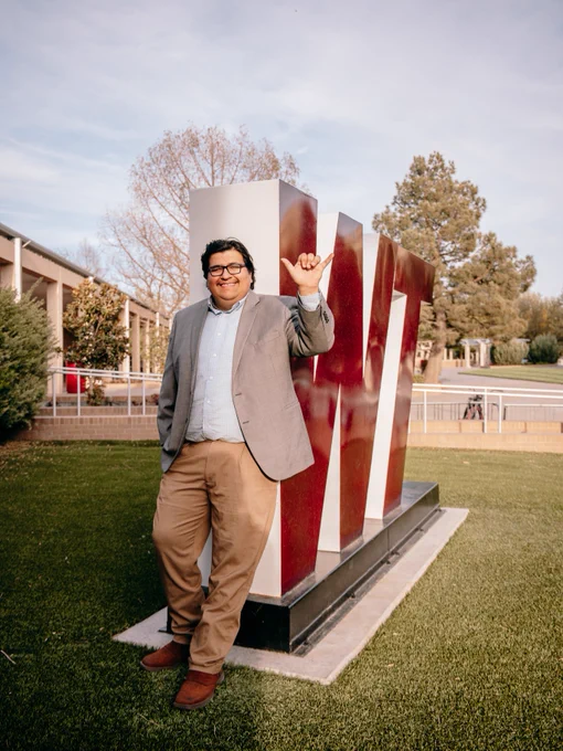 A person in a blazer and trousers stands smiling and making a gesture with one hand. They are standing next to a large, maroon "WT" sculpture on a grassy area, with trees and buildings in the background.

