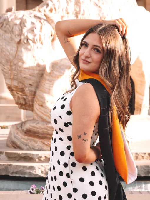 Young lady with brown hair posing in front of a marble buffalo statue. The lady is turned sideways with one hand behind her back and the other hand grabbing the hair on the top of her head. She is wearing a white dress with black polka dots and a black and yellow graduation hood around her neck. 