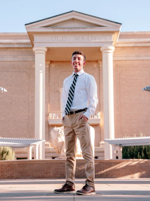 A person stands confidently in front of a building labeled "OLD MAIN," wearing a white shirt, striped tie, khaki pants, and brown shoes. The structure features classical columns and brick architecture. The scene is bright and clear.