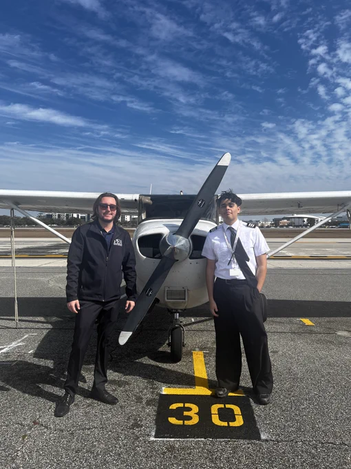 A small aircraft is parked on a tarmac. Two individuals in uniform are standing in front of the plane, smiling. The sky is clear with scattered clouds above.