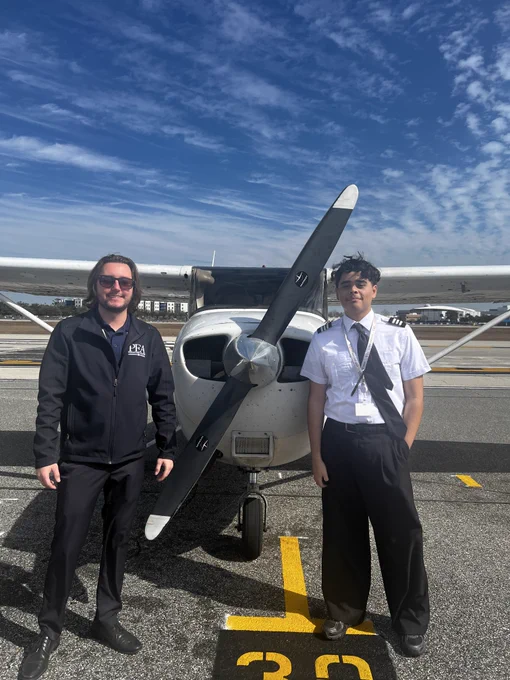Two people are standing in front of a small propeller airplane on a clear day. The runway and airport buildings are visible in the background.