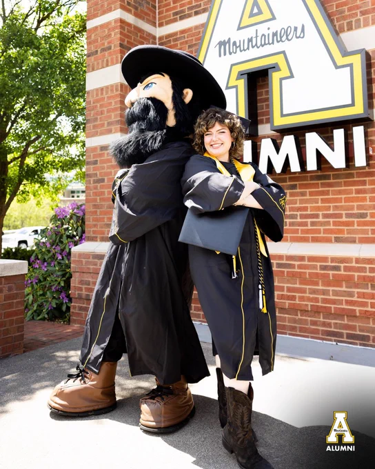 Graduate in a black cap and gown hugs Yosef in front of a large App State “Mountaineers” A display outside the McKinney Alumni Center. The graduate is smiling while holding a diploma cover and wearing black cowboy boots.