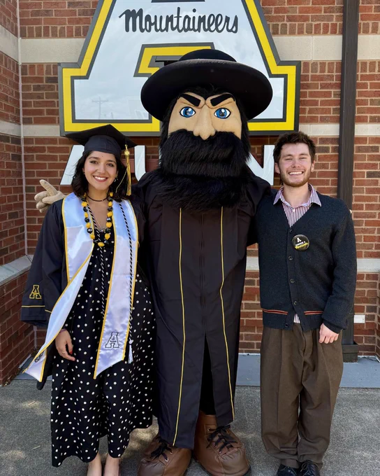Graduate in a black cap and gown with a white stole poses beside Yosef and another attendee in front of a large App State “Mountaineers” A display outside the McKinney Alumni Center.