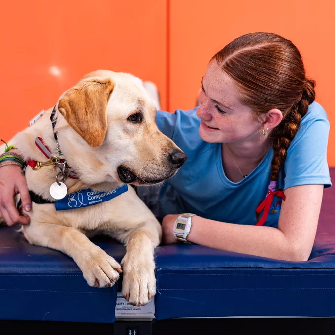 A young girl with braided hair lying on a mat, gently holding a calm yellow Labrador wearing a blue service dog vest.