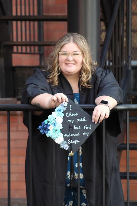student leaning on railing, holding grad cap that says "I chose my path, but God ordered my steps."