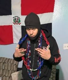 Image from court documents shows a photo of Westyn Lantigua in front of the flag of the Dominican Republic, throwing the Trinitario gang signs with his hands, while wearing a Trinitario beaded necklace. 