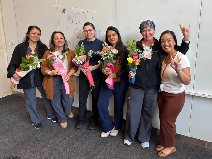 A group of six people standing indoors, each holding a bouquet of flowers. They are positioned in front of a whiteboard with "Happy Nurses Week" written on it. 