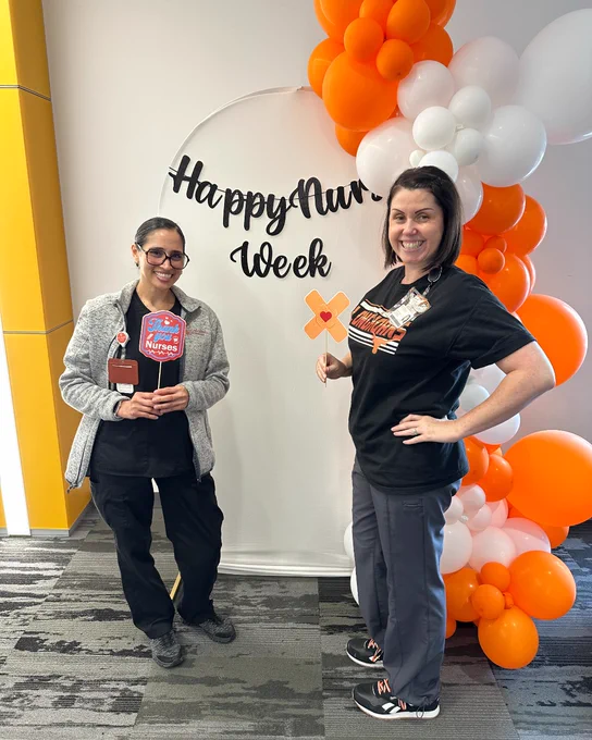 Two people stand smiling next to a "Happy Nurses Week" sign, surrounded by orange and white balloons.