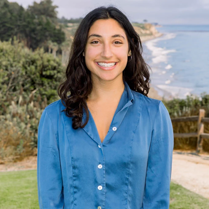A person in a blue blouse who is outdoors with a scenic background of lush greenery, a path, and a coastline with the ocean.