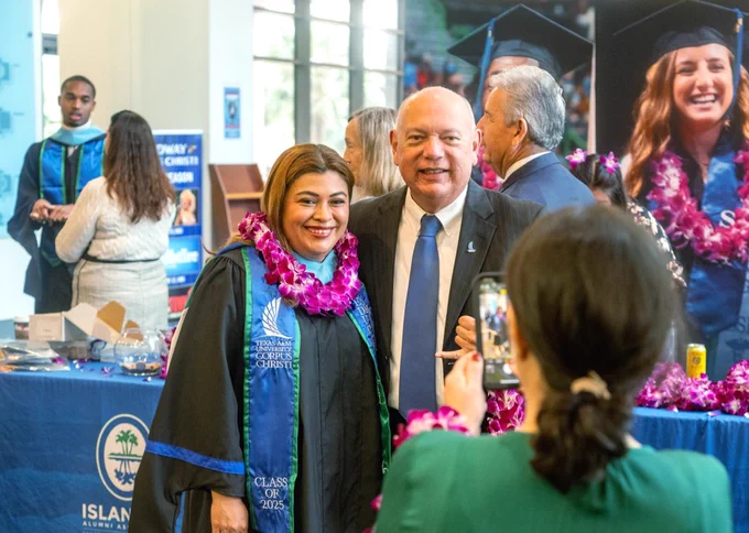 A Texas A&M University-Corpus Christi student in graduation attire with a lei stands next to another individual in formal wear, both smiling for a photo. They are indoors at an Island University TAMU-CC event, with a display in the background. Other attendees are visible in the area.