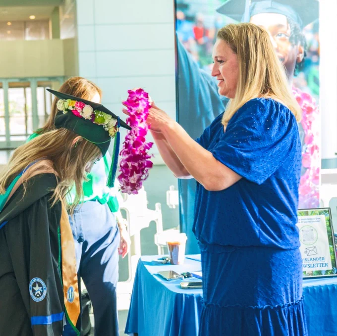 A Texas A&M University-Corpus Christi graduate in a graduation cap and gown receives a flower lei from a TAMU-CC alumna in a blue dress. A table with a sign that reads "Islander Newsletter" is visible nearby.