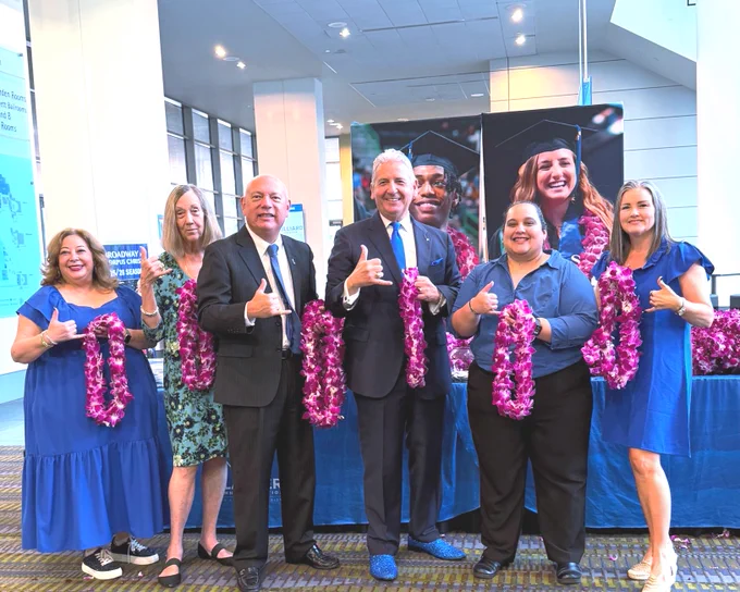 A group of Texas A&M University-Corpus Christi alumni stand together indoors on a patterned carpet. Most are holding purple flower leis. The group is gives skakas-up gestures and smiles, with a banner in the background showing TAMU-CC graduates in caps and gowns. Several are dressed in blue clothing.