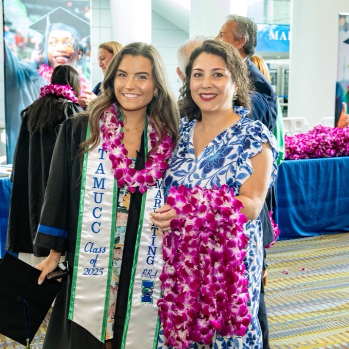A Texas A&M University-Corpus Christi alumna and graduate wearing a graduation gown with a "TAMUCC Class of 2025" stole and the other in a floral dress. Both hold purple leis. A graduation ceremony is taking place in the background.