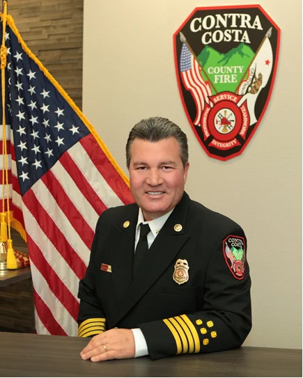 Contra Costa County Fire Protection District Fire Chief Aaron McAlister in dress uniform, seated at a desk with an American flag and the Con Fire shield logo on the wall behind him.