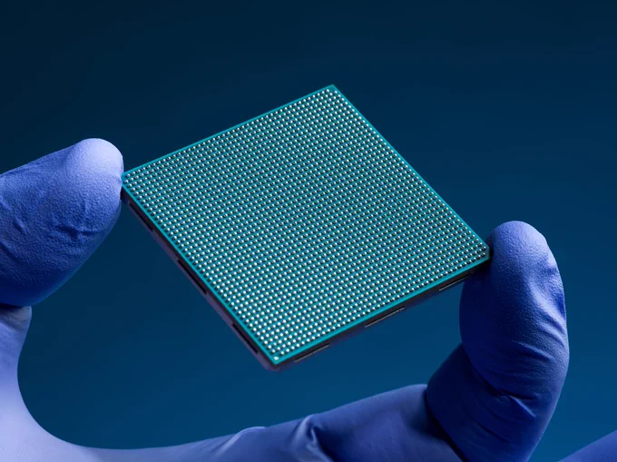 The finger and thumb of a hand clad in a blue nitrile glove hold a teal-colored square semiconductor chip against a dark blue background. The chip has an intricate grid of hundreds of tiny silver solder bumps on its underside.