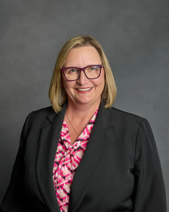 Headshot of Dr. Morris, a white woman with short blond hair and glasses. She wears a bright pink blouse and a dark gray suit jacket.