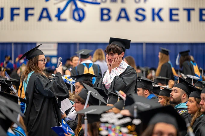 A graduation ceremony with graduates wearing black gowns and caps. A student is taking a photo with their smartphone.