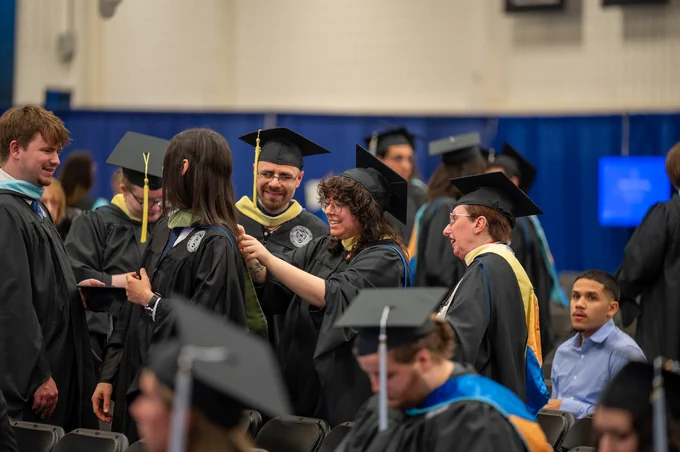 A graduation scene with graduates in black gowns and caps, some adjusting each other's stoles. A person in glasses smiles, observing the interaction. Others are seated, with varying expressions, in a brightly lit indoor space.