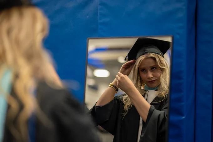 A person in a graduation gown and cap adjusts her cap while looking in a mirror. 