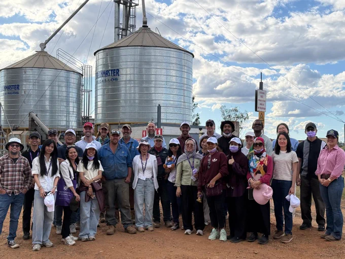 A group photo of participants and site experts standing at a feedlot staff with academics smiling in front of grain silos outside under a cloudy sky.