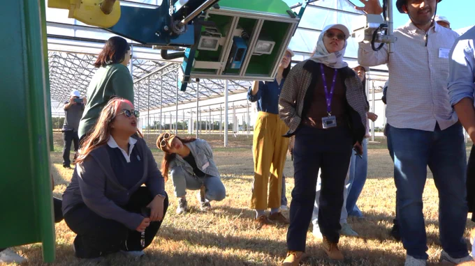 Participants crouching and standing to see agriculture technology and sensors on the extended arm of a tractor, in front of greenhouses
