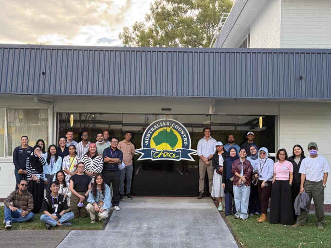 A group photo of participants smiling whilst with staff standing on either side of automatic doors with the Australian Country Choice logo in the centre. 
