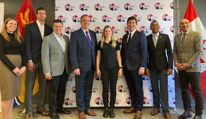 A group of people standing and smiling in front of a wall with "Research NB" logos. They are flanked by flags, including the Canadian flag.