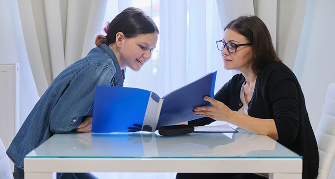 Two women sit at a table. One is holding and showing a blue folder to the other, who is leaning slightly forward. They appear to be having a discussion.