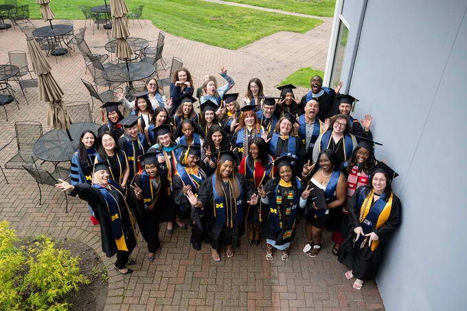 A group of graduates in caps and gowns pose for a celebratory photo on a patio. They are smiling and waving, with some wearing University of Michigan stoles. Tables, chairs, and a grassy area are visible in the background.