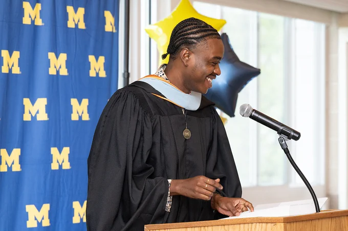 A person in a graduation gown stands at a podium, smiling and speaking into a microphone. The backdrop features the University of Michigan logo and there are blue and yellow star-shaped balloons in the background.
