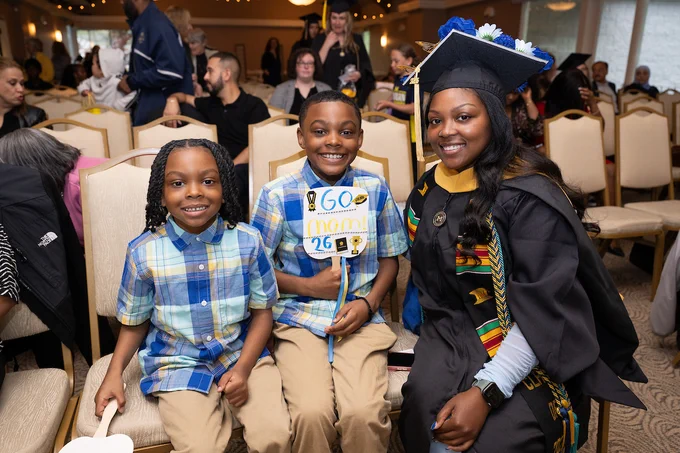A person in graduation attire sits smiling with two children beside them. One child holds a sign that reads "Go Mom! '26." There are several people seated in the background.