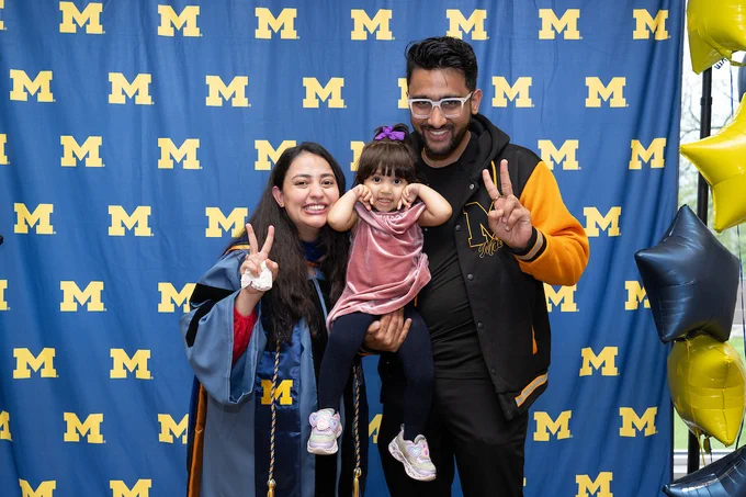 A group of three people smiling in front of a University of Michigan backdrop. Two adults stand on either side of a child, all making peace signs. Blue and yellow balloons are partially visible on the right.