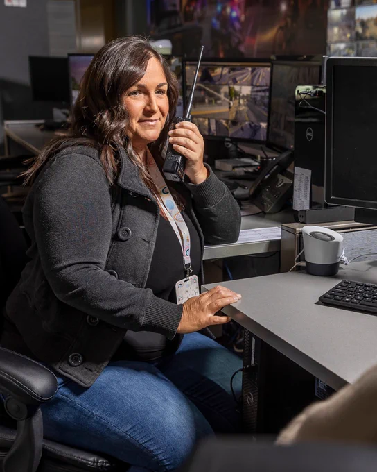 Woman security operator at a monitoring desk speaking into a radio, with multiple surveillance screens behind her.