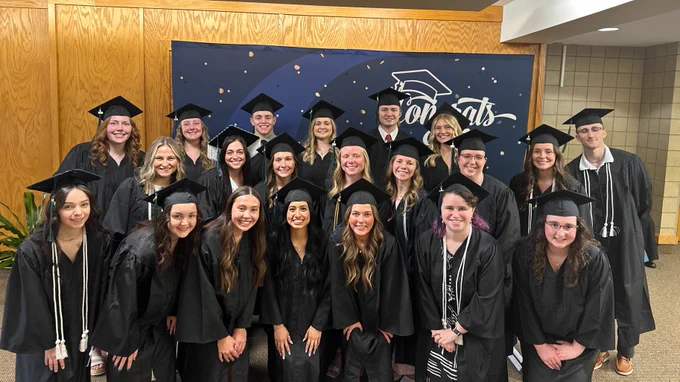 A group of people in graduation attire, wearing caps and gowns, stand in several rows. They are indoors, and behind them is a congratulatory banner. Everyone is smiling, celebrating their achievement.