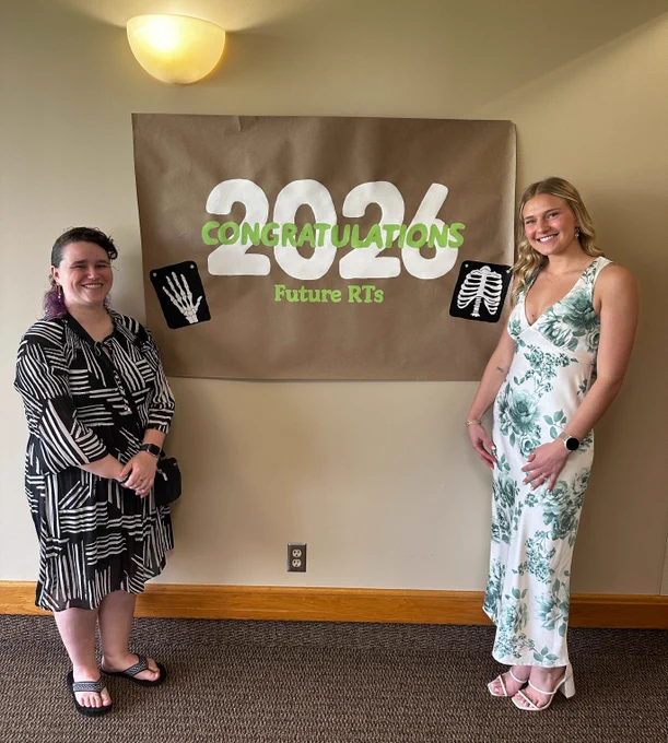 Two people are standing in front of a sign that reads "Congratulations 2026 Future RTs" with images of a hand X-ray and a rib X-ray. They are smiling and dressed in patterned outfits.