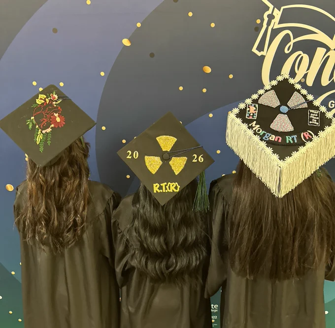 Three individuals in graduation gowns are standing with their backs to the camera. Their caps are decorated: one with a floral design, the second with nuclear symbols and the text "RITR" and "2026," and the third with patterns, symbols, and the words "Morgan RT.