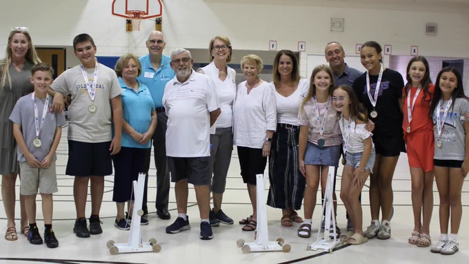 A group of people, including adults and children, stand in a gymnasium. Some children are wearing medals. They are posing in front of a few small, white wooden structures. There's a basketball hoop in the background.