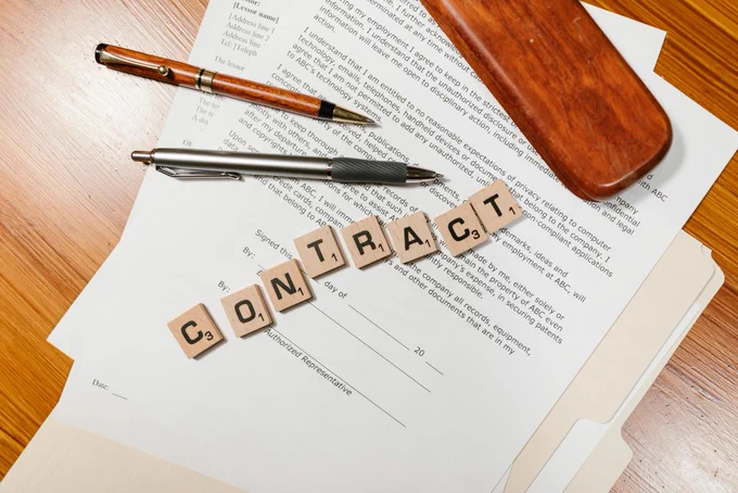 Letter tiles spelling out the word contract across some paperwork next to two pens.