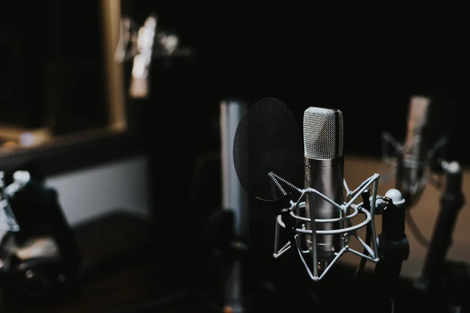 Studio condenser microphone in a shock mount with a pop filter, positioned in a dimly lit recording booth with a softly blurred background and warm, low-key lighting.