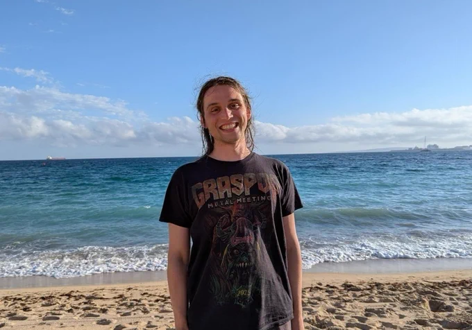 A photo of me smiling broadly at the camera. I am standing on a sandy beach, and right behind me are the beautiful blue waves of the Balearic Sea stretching out to the horizon under a bright, sunny sky.